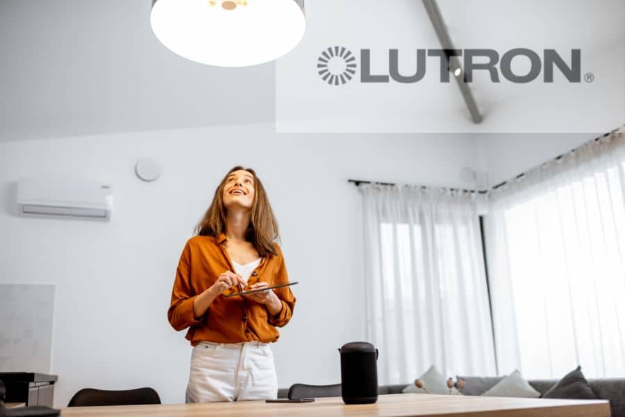 Woman standing underneath a light in her home, controlling it from her tablet with a Lutron logo in the upper right corner.